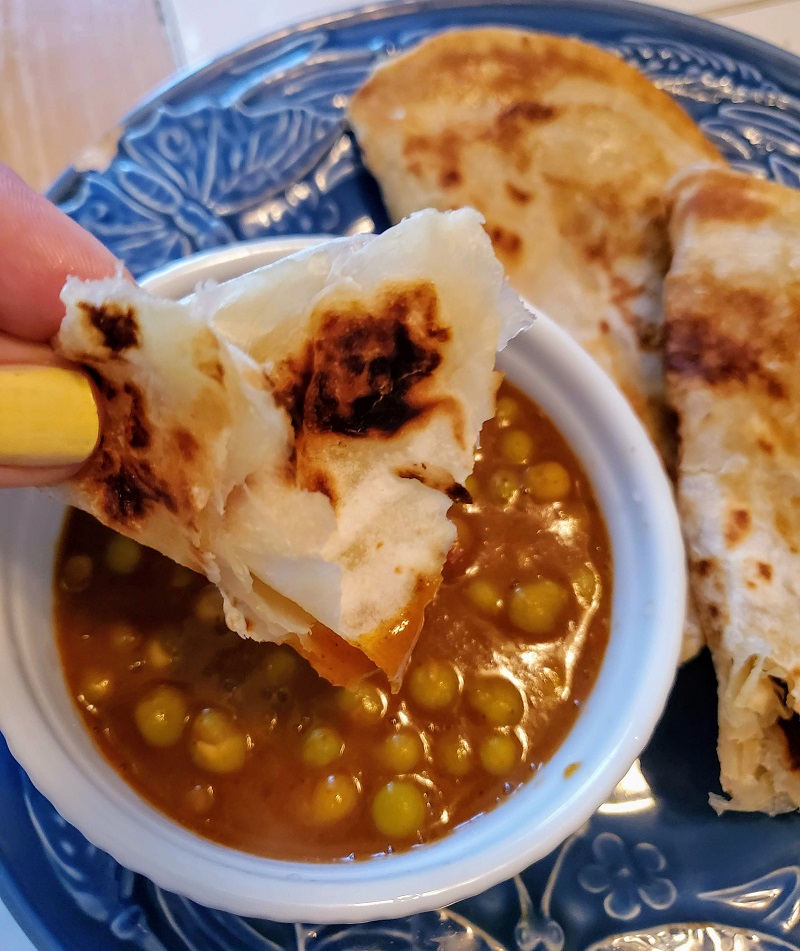 closeup of vindaloo pigeon peas in a white bowl and hand dipping a piece of paratha.