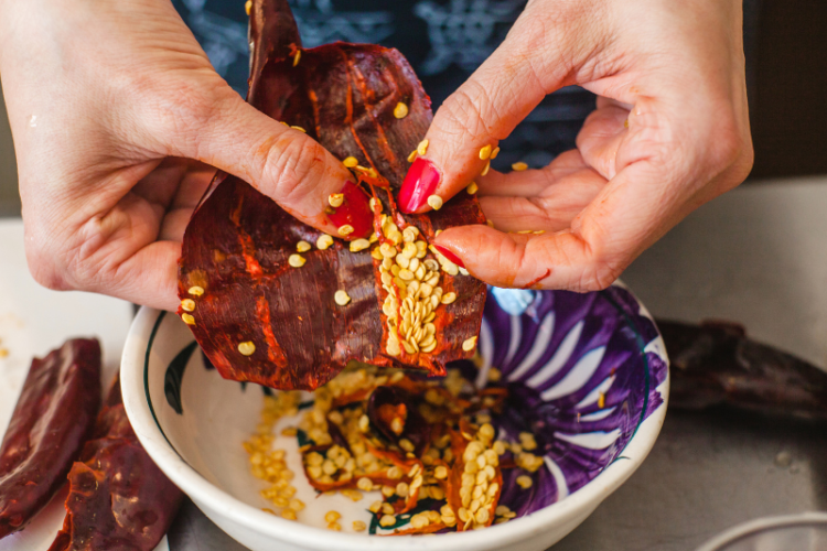 closeup of woman's hands de-seeding a dried chile pepper over bowl